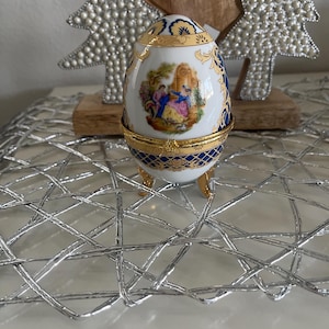 May include: A decorative porcelain egg with a hinged lid, featuring a painted scene of figures in a garden. The egg is white with gold and blue accents, and rests on small gold feet. A luxury collectible.