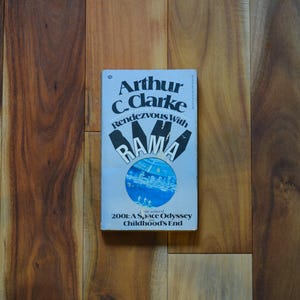 May include: A vintage paperback book titled "Rendezvous with Rama" by Arthur C. Clarke. The cover is light blue with black and white text and a circular illustration. The book is set against a wooden background.
