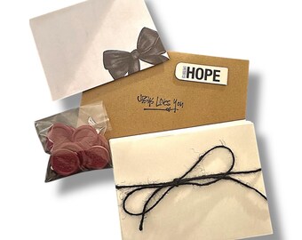 Puede incluir: Un surtido de artículos de papelería, que incluye un sobre blanco con un lazo negro, una tarjeta marrón con el texto "Jesus Loves You" y una pegatina "HOPE", y una pila de tarjetas blancas atadas con hilo negro. También se incluye una pequeña bolsa de lacres rojos.