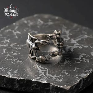 以下が含まれることがあります： 銀色のリングで、サソリのデザインです。リングは、爪と体がバンドを包み込む、詳細なサソリのデザインが特徴です。テクスチャ加工された濃い灰色の表面に表示されています。「Midnight 925 Craft」のブランドロゴが見えます。