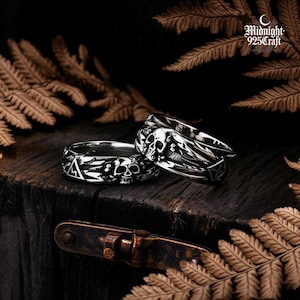 May include: Two silver rings with skull and geometric designs. One ring features a skull with wings, while the other displays a skull, triangle, and other symbols. The rings are on a dark wooden surface, with the text "Midnight 925Craft".