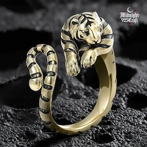May include: A gold-coloured ring with a detailed tiger design. The tiger's head and paws are sculpted, with black stripes. The ring wraps around the finger, creating a unique accessory. The ring is a statement piece, suitable for everyday wear.
