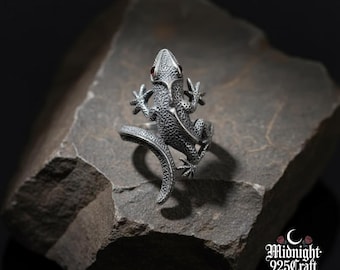 Puede incluir: Anillo plateado con forma de lagarto y ojos de piedras preciosas rojas. El anillo presenta escamas texturizadas y está colocado sobre una piedra gris. Una joya única que se enrolla alrededor del dedo.