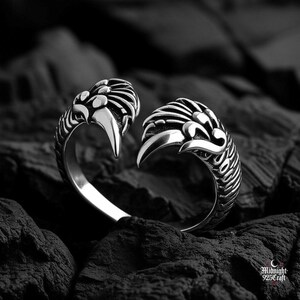 Gothic angel wing ring displayed on textured dark surface emphasizing intricate silverwork details. Each wing features multiple carved feathers with realistic depth and shadow. Adjustable sterling silver construction fits various finger sizes. Perfect gothic gift for dark angel fashion lovers.