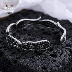 Side view of the sterling silver angel wings open cuff bangle displayed on a wet black lava stone surface. The bracelet's adjustable open-cuff design is visible, highlighting the tarnish-resistant 925 silver material and oxidized dark detailing. Perfect for everyday gothic or boho fashion wear.