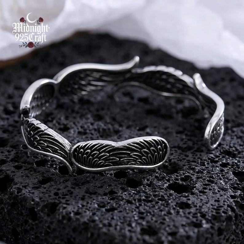 Side view of the sterling silver angel wings open cuff bangle displayed on a wet black lava stone surface. The bracelet's adjustable open-cuff design is visible, highlighting the tarnish-resistant 925 silver material and oxidized dark detailing. Perfect for everyday gothic or boho fashion wear.