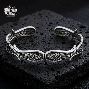 Close-up of an Angel Wings 925 Sterling Silver cuff bracelet resting on dark volcanic rock. The bracelet features intricately engraved feathered wings with an oxidized retro finish, showcasing the handcrafted Thai silver craftsmanship. Ideal as a gothic jewelry gift for her or a unique statement accessory.