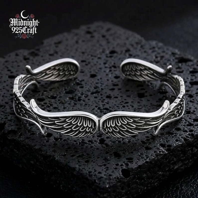 Close-up of an Angel Wings 925 Sterling Silver cuff bracelet resting on dark volcanic rock. The bracelet features intricately engraved feathered wings with an oxidized retro finish, showcasing the handcrafted Thai silver craftsmanship. Ideal as a gothic jewelry gift for her or a unique statement accessory.