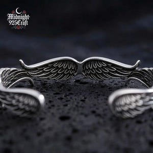 The Angel Wings sterling silver bracelet displayed on a black stone surface with moody atmospheric lighting, reflecting the dark gothic aesthetic of the jewelry. The 925 sterling silver shines against the dark background, perfect for a statement cuff bracelet gift or daily wear.