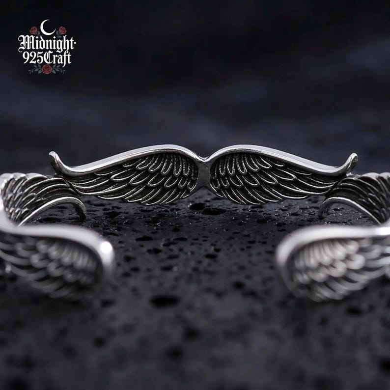 The Angel Wings sterling silver bracelet displayed on a black stone surface with moody atmospheric lighting, reflecting the dark gothic aesthetic of the jewelry. The 925 sterling silver shines against the dark background, perfect for a statement cuff bracelet gift or daily wear.