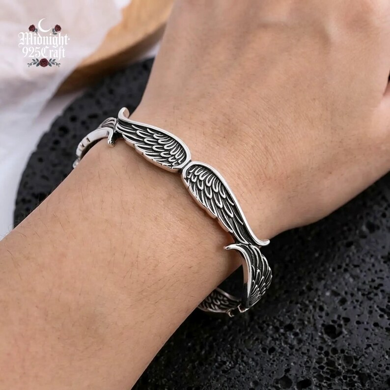 May include: A silver-colored bracelet with a wing design. The bracelet features detailed feather patterns and is worn on a wrist. The brand name "Midnight 925 Craft" is visible in the upper left corner.