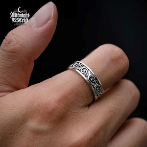 May include: A silver ring featuring an elaborate, swirling design. The ring is displayed on a finger, emphasizing the detailed pattern. The ring's surface reflects light, accentuating the intricate scrollwork. The ring is a piece of jewellery.