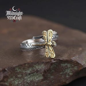 May include: A silver ring featuring a gold dragonfly design. The dragonfly has intricate wings and is the focal point of the ring. The ring band has a feather pattern. The text "Midnight 925 Craft" is in the upper left corner.