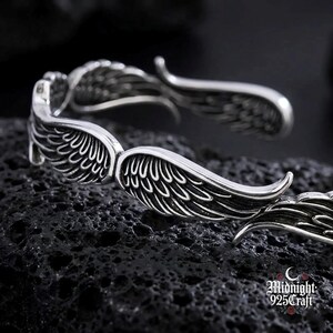 May include: A silver-colored bracelet with a detailed wing design. The bracelet features two sets of wings, with each wing having intricate feather detailing. The wings curve around the wrist, creating a unique and stylish accessory. The bracelet is displayed on a dark, textured surface.