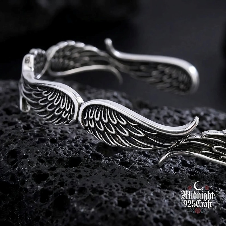 May include: A silver-colored bracelet with a detailed wing design. The bracelet features two sets of wings, with each wing having intricate feather detailing. The wings curve around the wrist, creating a unique and stylish accessory. The bracelet is displayed on a dark, textured surface.