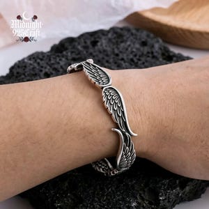 Lifestyle flat lay of the angel wing silver cuff bangle alongside a dark moody background. The bracelet's dark oxidized finish and retro Thai silver texture are prominent, making it an ideal gothic silver cuff for alternative fashion lovers, collectors, and unique jewelry seekers.