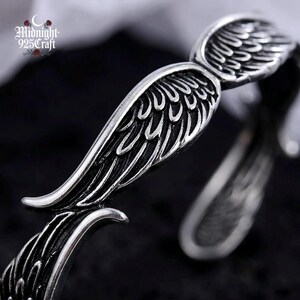 Pair of angel wing 925 sterling silver cuff bangles arranged side by side on volcanic rock, suggesting their use as couple matching jewelry. The oxidized finish and open cuff design are clearly visible, illustrating the adjustable fit and handmade artisan quality.