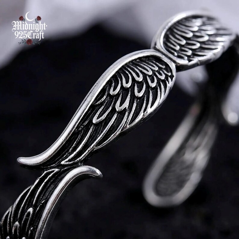 Pair of angel wing 925 sterling silver cuff bangles arranged side by side on volcanic rock, suggesting their use as couple matching jewelry. The oxidized finish and open cuff design are clearly visible, illustrating the adjustable fit and handmade artisan quality.