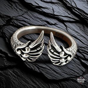 Close-up view of sterling silver gothic angel wing ring showing detailed feather texture and craftsmanship. Adjustable 925 silver band with dual wing design, each feather individually carved. Dark moody photography on weathered wood surface highlights intricate silverwork and gothic aesthetic.