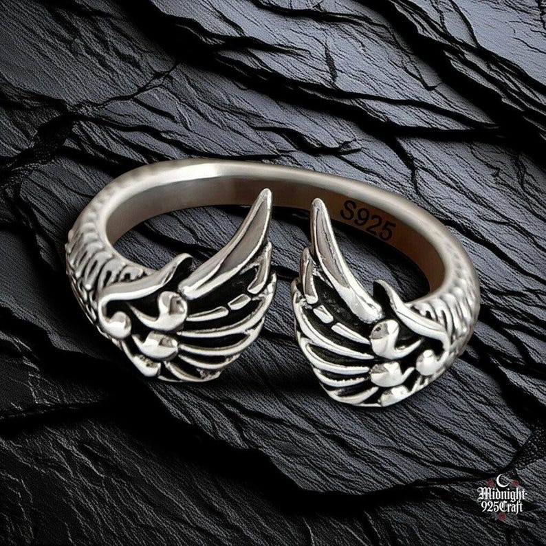 Close-up view of sterling silver gothic angel wing ring showing detailed feather texture and craftsmanship. Adjustable 925 silver band with dual wing design, each feather individually carved. Dark moody photography on weathered wood surface highlights intricate silverwork and gothic aesthetic.
