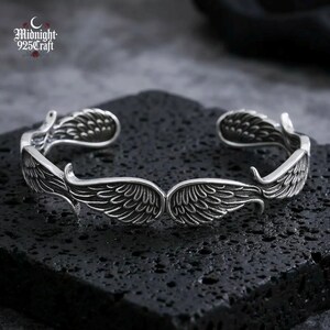May include: A silver-colored cuff bracelet with an open design. The bracelet features detailed wing designs on each end, with the text "Midnight 925 Craft" in the upper left corner. The bracelet is displayed on a textured black surface.