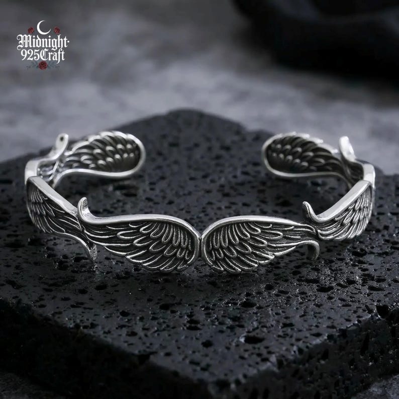 May include: A silver-colored cuff bracelet with an open design. The bracelet features detailed wing designs on each end, with the text "Midnight 925 Craft" in the upper left corner. The bracelet is displayed on a textured black surface.