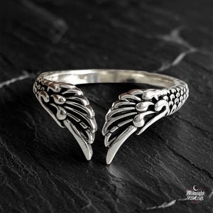Puede incluir: Un anillo plateado con un diseño de alas de dragón. El anillo presenta dos motivos detallados de alas de dragón que parecen envolver el dedo. El anillo está sobre una superficie oscura y texturizada, resaltando los intrincados detalles de la joya.