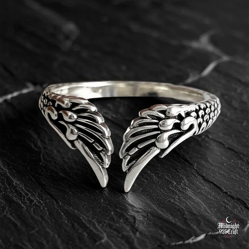 Gothic angel wing ring in 925 sterling silver with intricate feather detailing, adjustable band, photographed on dark textured wood background. Features dual angel wings with detailed carved feathers wrapping around adjustable silver band. Perfect for goth fashion, dark angel aesthetic, alternative jewelry lovers.