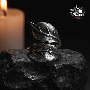 Op de afbeelding: Een zilverkleurige ring in de vorm van twee overlappende bladeren. De ring staat tegen een donkere achtergrond, met een brandende kaars en de tekst "Midnight 925Craft" zichtbaar. Het ontwerp van de ring heeft gedetailleerde adering.