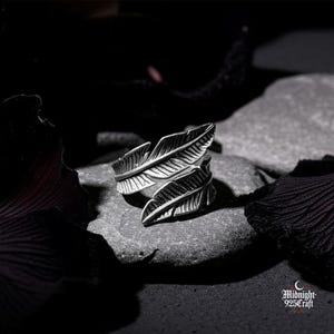 Puede incluir: Un anillo plateado con diseño de plumas. El anillo presenta dos plumas estilizadas que se envuelven alrededor del dedo. El anillo está sobre un fondo oscuro con el logo "Midnight 925 Craft".