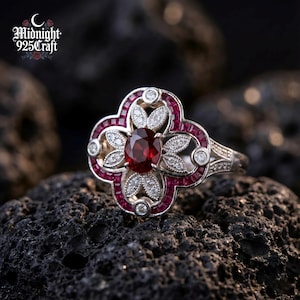Puede incluir: Un anillo de plata con un diseño de trébol de cuatro hojas, con una gran gema roja ovalada en el centro, rodeada de piedras rojas más pequeñas y piedras de acento transparentes. El anillo está sobre una superficie oscura y texturizada. El texto "Midnight 925 Craft" es visible.