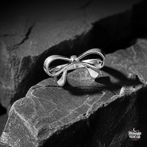 Può includere: Un anello color argento a forma di fiocco. L'anello è posto su uno sfondo scuro e strutturato. Il design del fiocco è dettagliato ed elegante, con una finitura lucida. È un gioiello.