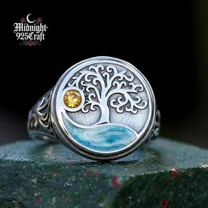 Op de afbeelding: Zilveren ring met een gedetailleerd boomontwerp in een cirkelvormige lijst. De ring heeft een gele edelsteen en een blauwe emaille basis. De ring is gestempeld met "Midnight 925 Craft".
