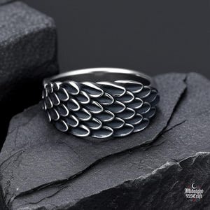 Peut inclure: Bague en argent avec un motif texturé en forme d'écailles. La partie texturée présente une finition sombre oxydée, contrastant avec le bandeau en argent poli. La bague est présentée sur une surface texturée gris foncé. Bijou.
