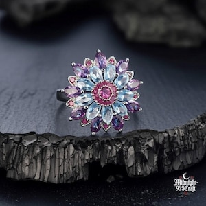 Pode incluir: Anel de prata com design floral e pedras preciosas multicoloridas. As pétalas são compostas por pedras rosa, azuis e roxas, com um centro rosa escuro. O anel é exibido sobre uma superfície escura e texturizada, realçando as cores.