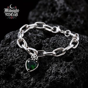 Op de afbeelding: Een zilveren kettingarmband met een hartvormige hanger. De hanger heeft een donkergroene edelsteen en een kruisontwerp. De armband wordt getoond op een donkere, getextureerde ondergrond. De merknaam "Midnight 925 Craft" is zichtbaar.