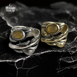 Op de afbeelding: Twee ringen met veermotieven en ovale gele edelstenen. Eén ring is zilverkleurig, de andere goudkleurig. De ringen hebben de tekst "Midnight 925 Craft" in de linkerbovenhoek.