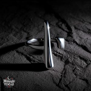 Op de afbeelding: Een zilveren ring met een modern ontwerp. De ring heeft een verticale staaf en een kleine, rechthoekige accent. De ring is geplaatst tegen een donkere, getextureerde achtergrond. Het Midnight 925 Craft logo is linksonder zichtbaar.