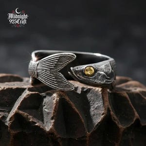 May include: A silver-colored ring in the shape of a fish. The ring features a fish tail and head design with a gold-colored eye. The ring is displayed on a textured, dark brown surface. The brand name "Midnight 925 Craft" is visible.