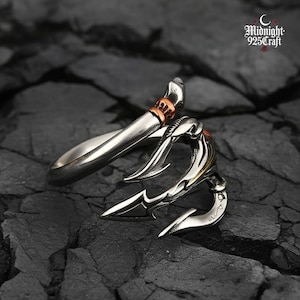 Puede incluir: Un anillo plateado con un diseño de tridente. El anillo presenta una cabeza de tridente detallada con tres púas y una banda decorativa. El anillo tiene un acento de color cobre y está sobre un fondo oscuro y texturizado.