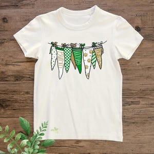 May include: White t-shirt featuring a string of pennant flags in green, gold, and white. The flags display patterns and the words "Burton" and "Wolves". The shirt has a crew neck and short sleeves, perfect for casual wear.