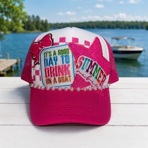 May include: A hot pink trucker hat with a white mesh back and a curved brim. The hat features a pink star with the word "SUMMER" and a patch that says "IT'S A GOOD DAY TO DRINK ON A BOAT". A string of pearls adorns the brim.