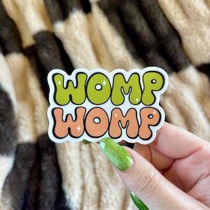 May include: A sticker with the word "WOMP" in green and orange bubble letters. The sticker is held by a hand with green glitter nail polish. The background is a brown, black, and white patterned fabric.