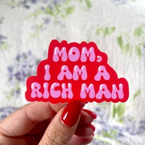 May include: A red sticker with the text "MOM, I AM A RICH MAN" in pink bubble letters. The sticker is held in a hand with red nail polish against a floral background.