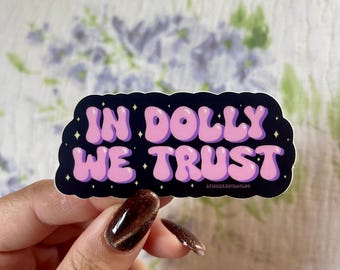 May include: A sticker featuring the text "IN DOLLY WE TRUST" in pink bubble letters with a purple outline, set against a black background with white stars. The sticker has an irregular, wavy shape.