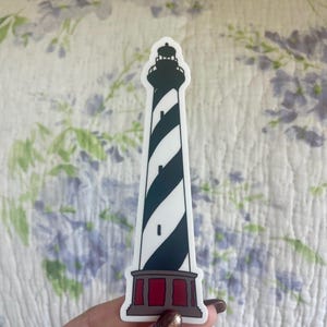 Puede incluir: Una pegatina con un faro con una torre a rayas blancas y negras, una parte superior negra y una base roja. El diseño del faro es una pegatina, probablemente para un coche, un portátil o una botella de agua.