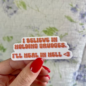 Peut inclure: Autocollant blanc avec texte orange : « I BELIEVE IN HOLDING GRUDGES. I'LL HEAL IN HELL <3 ». L'autocollant a une police rétro et est sur un fond floral.