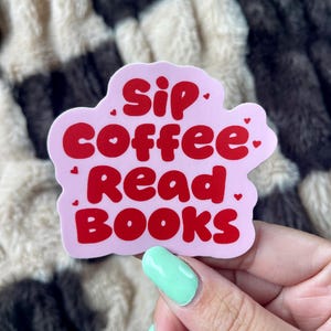May include: A die-cut sticker with a light pink background and red text that reads Sip coffee Read Books, accented with small red hearts. The sticker has a wavy, irregular border and features bold, rounded typography for the lettering.