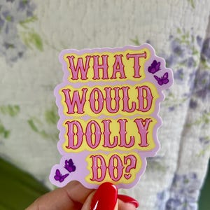 Pode incluir: Um autocolante com o texto "WHAT WOULD DOLLY DO?" em rosa e amarelo, com detalhes de borboletas roxas. O autocolante tem um fundo lilás claro. O autocolante é segurado por uma pessoa.