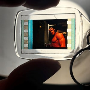 May include: A clear plastic keychain featuring a film strip with a scene from the movie Hellboy. The image shows the character Hellboy with red skin and horns. The keychain has a silver key ring.